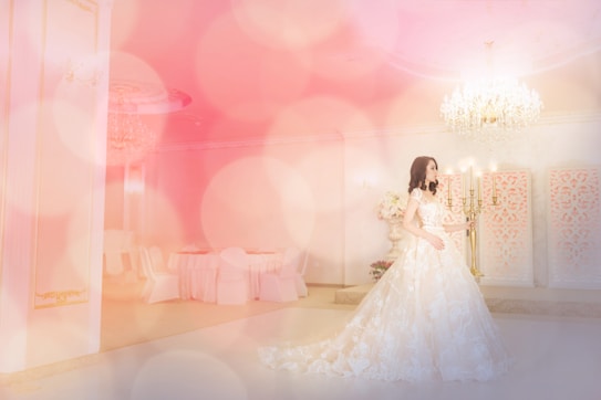 A woman wearing a long, elegant bridal gown in an opulent setting with soft, ambient lighting and a large chandelier. The room features decorative panels on the walls, a lavish candelabra, and a round table with chairs covered in white fabric creating a luxurious and dreamy atmosphere.