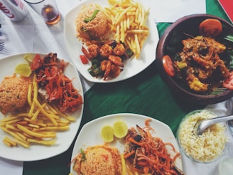 A variety of dishes are arranged on a table, including plates with rice, grilled vegetables, fried potatoes, and meats. Some plates hold shrimp with sautéed vegetables and sauces, while another has a curry in a wooden bowl garnished with tomato slices. A small bowl with white rice and a serving spoon is also visible along with a can of soda and a glass with a dark liquid.