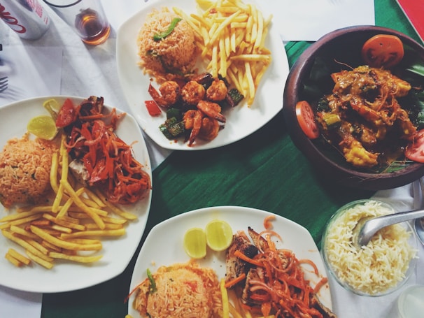 A variety of dishes are arranged on a table, including plates with rice, grilled vegetables, fried potatoes, and meats. Some plates hold shrimp with sautéed vegetables and sauces, while another has a curry in a wooden bowl garnished with tomato slices. A small bowl with white rice and a serving spoon is also visible along with a can of soda and a glass with a dark liquid.