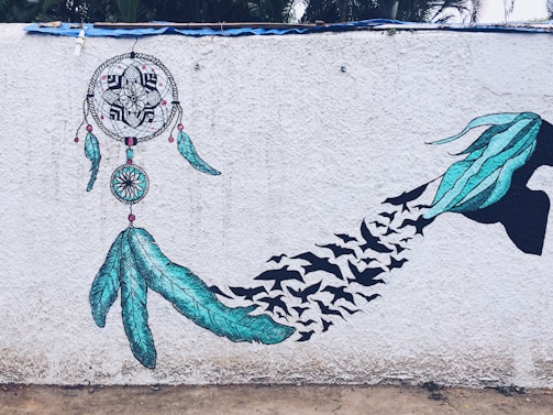 A mural depicting a dreamcatcher with intricate designs and two large blue feathers, transitioning into a silhouette of a hand releasing a flock of birds. The artwork is set against a textured white wall with green plants visible at the top.