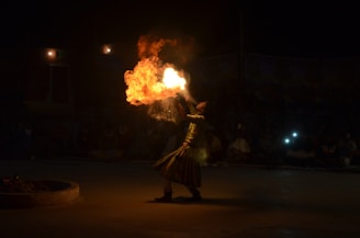 A performer engaging in a fire-breathing act, surrounded by darkness with an audience barely visible in the background. The flame is bright and fiery, creating a dramatic contrast against the dimly lit environment.