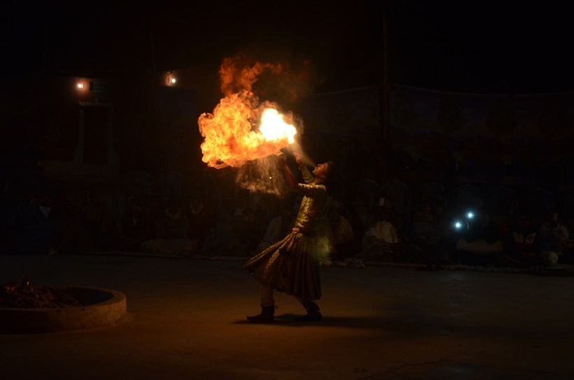 A performer engaging in a fire-breathing act, surrounded by darkness with an audience barely visible in the background. The flame is bright and fiery, creating a dramatic contrast against the dimly lit environment.