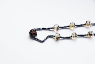 A beaded necklace featuring interconnected strands of small, dark beads interspersed with larger, transparent beads. The necklace is crafted with a prominent flower-shaped bead on one end, adding an artistic flair. The arrangement is displayed on a plain white background, highlighting the simplicity and elegance of the piece.