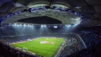 An expansive soccer stadium filled with spectators, illuminated by bright overhead lights, showcasing a well-maintained green field with players scattered across it. The stadium's curved roof and seating areas create a grand and dramatic atmosphere.