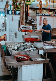 A bustling market scene with a variety of seafood displayed on metal tables. A man in a blue shirt is standing behind the counter, possibly preparing or selling the products. Colorful decorations hang above the stand, and there are numbers and signs in the background.
