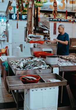 A bustling market scene with a variety of seafood displayed on metal tables. A man in a blue shirt is standing behind the counter, possibly preparing or selling the products. Colorful decorations hang above the stand, and there are numbers and signs in the background.