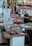 A bustling market scene with a variety of seafood displayed on metal tables. A man in a blue shirt is standing behind the counter, possibly preparing or selling the products. Colorful decorations hang above the stand, and there are numbers and signs in the background.