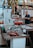 A bustling market scene with a variety of seafood displayed on metal tables. A man in a blue shirt is standing behind the counter, possibly preparing or selling the products. Colorful decorations hang above the stand, and there are numbers and signs in the background.