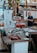 A bustling market scene with a variety of seafood displayed on metal tables. A man in a blue shirt is standing behind the counter, possibly preparing or selling the products. Colorful decorations hang above the stand, and there are numbers and signs in the background.