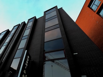 A modern high-rise building with a series of windows stretches upward towards a bright blue sky. The building's facade is constructed from dark bricks, while adjacent structures display orange-brown brickwork. The reflection of a crane is visible in one of the windows, adding an element of urban development.