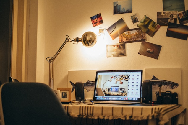 A cozy workspace with a wooden desk features an open laptop displaying a digital collage. A vintage desk lamp casts a warm glow, illuminating a wall adorned with various photographs. A digital camera and framed picture sit on the desk, creating a personal and creative atmosphere.