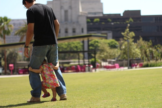 A man wearing a black T-shirt and jeans stands on a grassy field as a small child in a colorful dress clings to his legs. The background features modern buildings and trees, creating a city park setting with bright outdoor furniture visible in the distance.