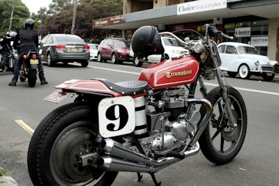 A classic motorbike parked on a street next to other vehicles, including a vintage car and several modern cars. The bike features a red fuel tank with the word 'Triumphant' and a racing number plate with the number 9. In the background, there are shops and people, with a person on another motorcycle.