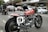A classic motorbike parked on a street next to other vehicles, including a vintage car and several modern cars. The bike features a red fuel tank with the word 'Triumphant' and a racing number plate with the number 9. In the background, there are shops and people, with a person on another motorcycle.
