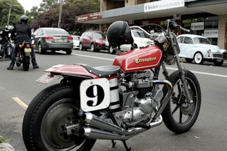 A classic motorbike parked on a street next to other vehicles, including a vintage car and several modern cars. The bike features a red fuel tank with the word 'Triumphant' and a racing number plate with the number 9. In the background, there are shops and people, with a person on another motorcycle.