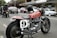 A classic motorbike parked on a street next to other vehicles, including a vintage car and several modern cars. The bike features a red fuel tank with the word 'Triumphant' and a racing number plate with the number 9. In the background, there are shops and people, with a person on another motorcycle.