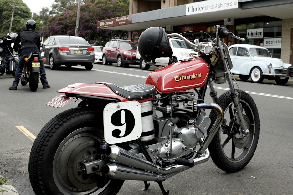 A classic motorbike parked on a street next to other vehicles, including a vintage car and several modern cars. The bike features a red fuel tank with the word 'Triumphant' and a racing number plate with the number 9. In the background, there are shops and people, with a person on another motorcycle.