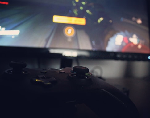 A close-up view of a gaming controller lying in front of a monitor displaying a video game. The scene is dimly lit, with the controller buttons faintly highlighted by the screen light. The screen image is somewhat blurred, suggesting focus on the foreground controller.
