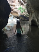 A person is wading through a shallow river in a narrow canyon. The rocky canyon walls tower on either side, with sunlight illuminating parts of the scene. In the background, two other people are seen standing on rocks beside the flowing water. Some greenery is visible amongst the rocks.
