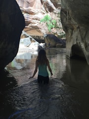 A person is wading through a shallow river in a narrow canyon. The rocky canyon walls tower on either side, with sunlight illuminating parts of the scene. In the background, two other people are seen standing on rocks beside the flowing water. Some greenery is visible amongst the rocks.
