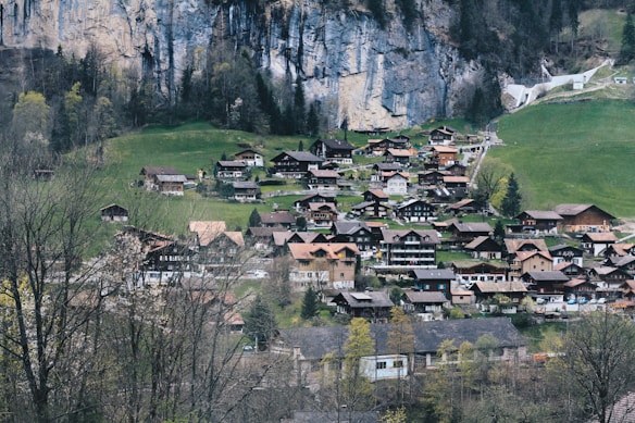 A picturesque village nestled in a lush, mountainous landscape with numerous charming wooden houses. The backdrop features a steep, rocky cliff with sparse trees, while the foreground is dominated by vibrant green grass and budding trees, suggesting early spring.