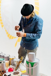 A person in a denim shirt and black hat concentrates on mixing paint in a small container. Several paint cans and brushes are scattered on the table, with one can having green paint dripping down the side. The background is a light color with yellow patterns.