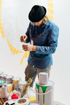 A person in a denim shirt and black hat concentrates on mixing paint in a small container. Several paint cans and brushes are scattered on the table, with one can having green paint dripping down the side. The background is a light color with yellow patterns.