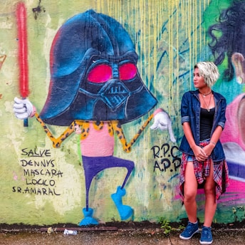 A vibrant mural features a cartoon character with a large helmet and bright red eyes, holding a glowing red stick. The character is colorful with blue, pink, and yellow leopard print details. Beside the mural, a person with light hair, wearing a denim jacket and plaid shirt tied around their waist, poses against the wall.