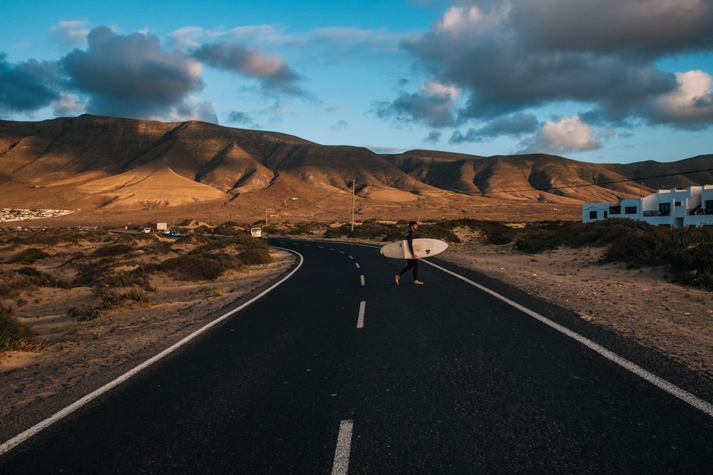 person carrying surfboard on asphalt road near brown mountains