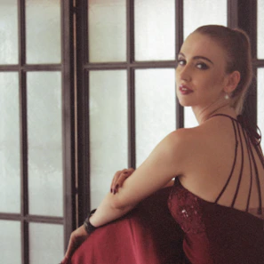 A sensual woman in an elegant red dress smiling softly in a cozy room.