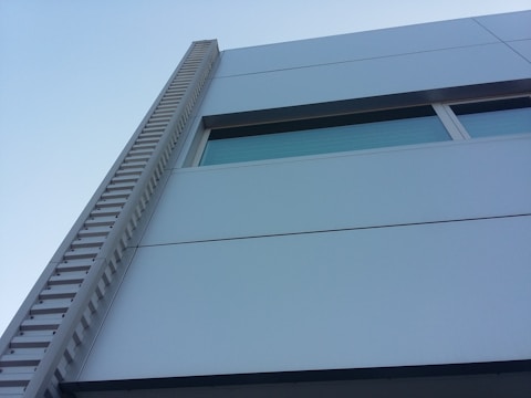 A modern building facade featuring a light gray exterior with clean horizontal lines and geometric shapes. A tall, ribbed architectural feature runs vertically along the side, and there is a window with a blue-tinted glass panel.