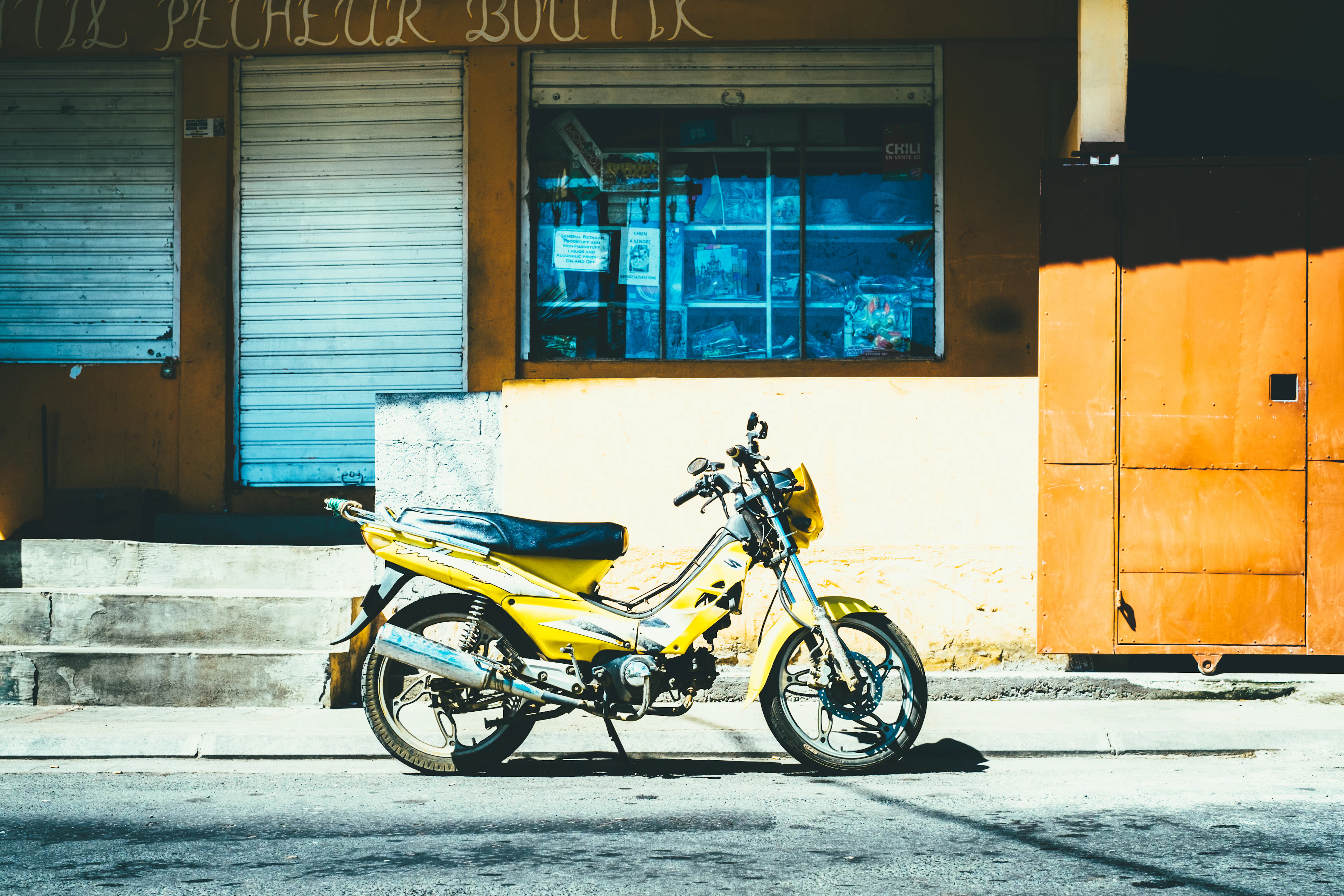 A yellow moped is parked outside a shuttered shopfront on a quiet sunlit street.