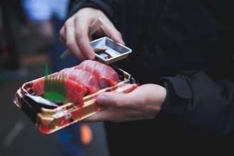 A person is holding a tray of fresh sushi with sliced pieces of raw fish, accompanied by a small container of soy sauce and chopsticks. The person is wearing a dark jacket and is outdoors, possibly in a marketplace or festival setting.