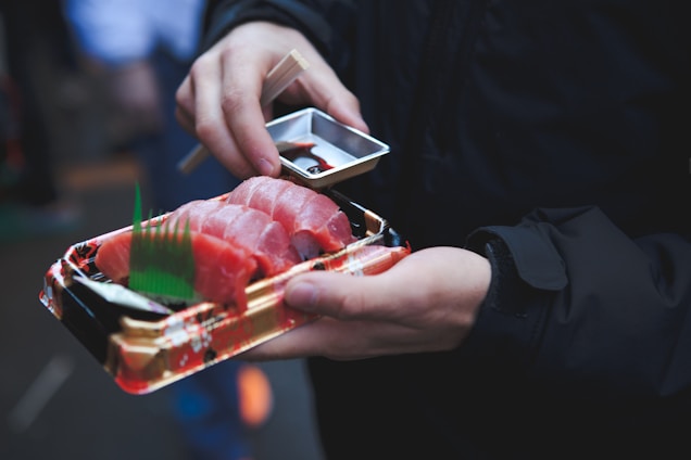 A person is holding a tray of fresh sushi with sliced pieces of raw fish, accompanied by a small container of soy sauce and chopsticks. The person is wearing a dark jacket and is outdoors, possibly in a marketplace or festival setting.