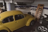 A vintage yellow car parked inside a garage with various items scattered around. The garage has white tiled walls with a blue stripe and contains objects such as car tires, a ladder leaning against the wall, and shelves with car maintenance supplies.