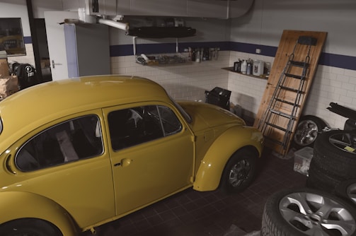 A vintage yellow car parked inside a garage with various items scattered around. The garage has white tiled walls with a blue stripe and contains objects such as car tires, a ladder leaning against the wall, and shelves with car maintenance supplies.