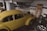 A vintage yellow car parked inside a garage with various items scattered around. The garage has white tiled walls with a blue stripe and contains objects such as car tires, a ladder leaning against the wall, and shelves with car maintenance supplies.