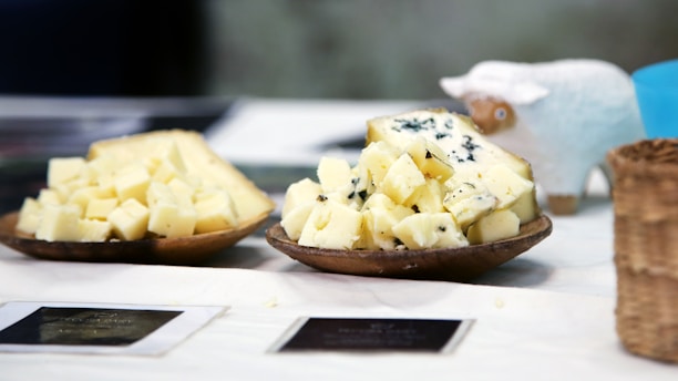 Two wooden plates hold chunks of cheese, one of which appears to be a blue cheese with visible blue veins. A small sheep figurine is placed behind the cheese, adding a whimsical touch. The setting includes a woven basket and what appears to be a label or sign, all on a white tablecloth.