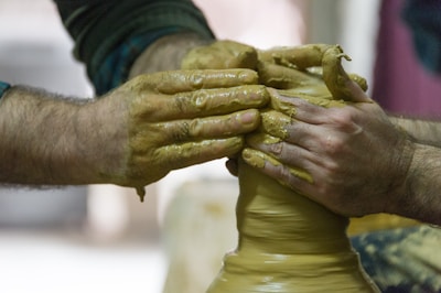 Two pairs of hands are shaping wet clay on a potter's wheel, with one pair providing steady support and the other actively molding the clay. The hands are covered in a smooth layer of clay, illustrating the tactile and hands-on nature of pottery.