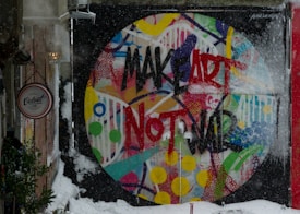 A vibrant, circular mural with the phrase 'MAKE ART NOT WAR' painted in bold black and red letters on a multicolored abstract background. The wall is in an outdoor wintry setting, partially covered with snow. A sign for 'Velvet Cafe' is visible to the side, with trees and snow around.