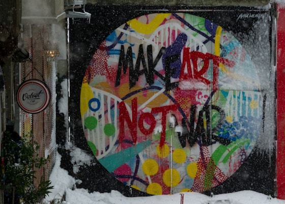 A vibrant, circular mural with the phrase 'MAKE ART NOT WAR' painted in bold black and red letters on a multicolored abstract background. The wall is in an outdoor wintry setting, partially covered with snow. A sign for 'Velvet Cafe' is visible to the side, with trees and snow around.