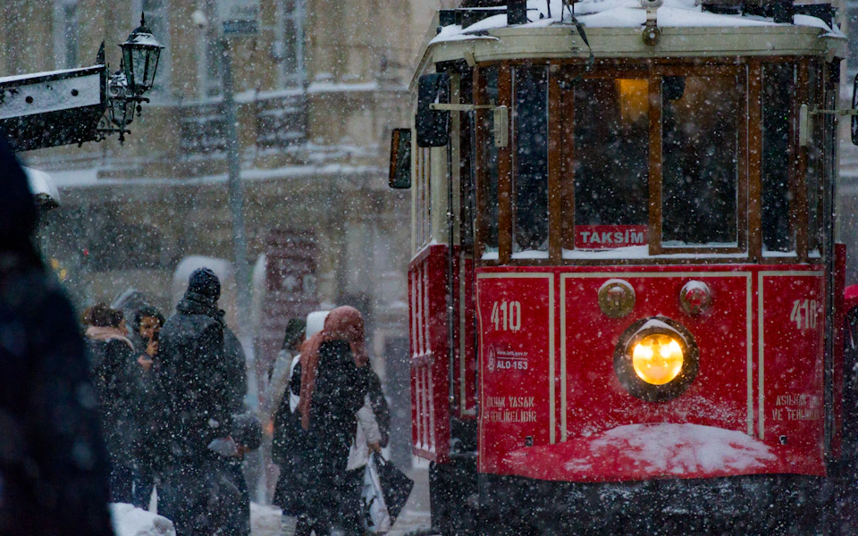 The red nostalgic tram on İstiklal Avenue, its headlight on and its sign reading Taksim, in heavy falling snow with people in winter coats beside it