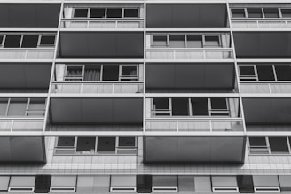 The image features the facade of a modern apartment building with a grid-like design. Each section comprises large windows and balconies framed by metallic structures. The architecture appears utilitarian and repetitive, creating a sense of order and symmetry.