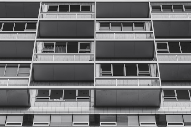 The image features the facade of a modern apartment building with a grid-like design. Each section comprises large windows and balconies framed by metallic structures. The architecture appears utilitarian and repetitive, creating a sense of order and symmetry.