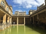 An ancient Roman-style bath structure with tall columns surrounding a large central pool of green water. The architecture features arches, detailed stone work, and statues atop the roofline. The sky is bright with scattered clouds, and several people are seen walking around and sitting near the water.
