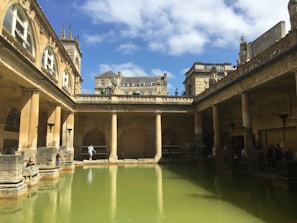 An ancient Roman-style bath structure with tall columns surrounding a large central pool of green water. The architecture features arches, detailed stone work, and statues atop the roofline. The sky is bright with scattered clouds, and several people are seen walking around and sitting near the water.