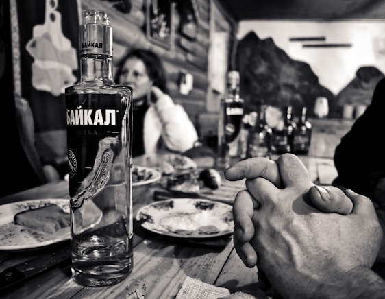 A monochrome image of a cozy indoor setting showing a close-up of a bottle labeled with Russian text, presumably vodka, on a wooden table. Nearby are plates with food remnants and clasped hands in the foreground. People sit around the table in a rustic environment, with blurred artwork on the walls.