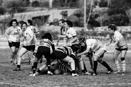 A group of women engaged in a rugby game on a grassy field, competing intensely as they grapple with each other for possession of the ball. The players are wearing numbered jerseys and some have protective gear like mouthguards and scrum caps. The background features blurred foliage and structures, indicating the setting is an outdoor sports field.