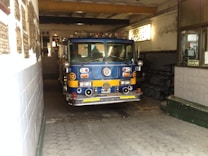 A vintage blue and yellow fire truck is parked inside a garage with a hexagonal tile floor. The garage interior has white and gray tiled walls and a weathered look with plaques on the walls and an exit sign visible. The fire truck, numbered 12, features a classic design and various equipment and lights.