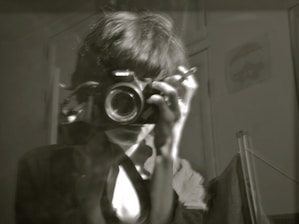 A black and white image of a person holding a camera up to their face, possibly taking a photograph. Their hair is slightly messy, and they are wearing a jacket over a shirt. The room is dimly lit, and the background shows part of a wall with a faint poster or picture.